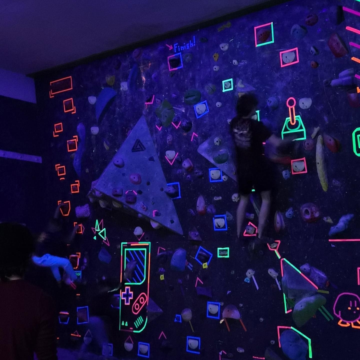 People climbing on an indoor rock wall with neon tape lit by blacklights at Shasta Rock Club in Redding, California.