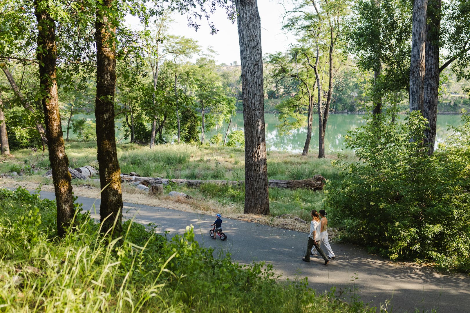 Two adults walk side by side while a child rides a small bike along a paved, tree-lined trail beside the Sacramento River in Redding, California. Sunlight filters through the trees, creating a calm and inviting outdoor scene.