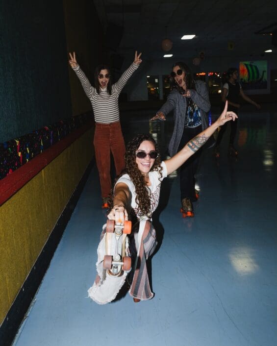 Three smiling women wearing sunglasses and striking fun poses while roller skating on the blue rink floor at Viking Skate Country in Redding. The Viking ship artwork is visible in the background as they enjoy their retro skating adventure.