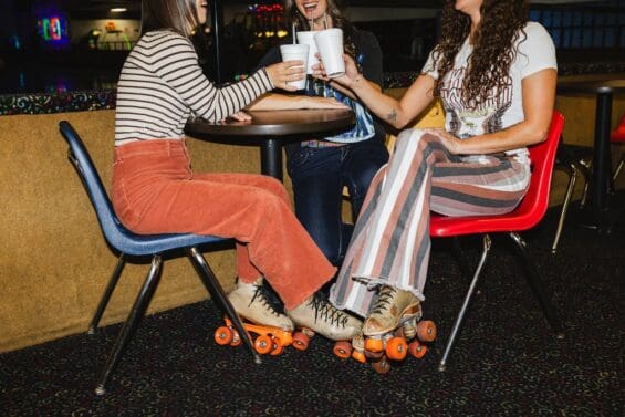 Three friends in roller skates toasting with drinks in white styrofoam cups while seated at a table in Viking Skate Country. They're wearing colorful retro pants with their skates still on, showcasing vintage roller rink vibes.