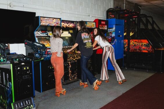 Three women on roller skates playing arcade games at Viking Skate Country in Redding. They're wearing colorful retro pants and enjoying the vintage arcade area of the roller rink.