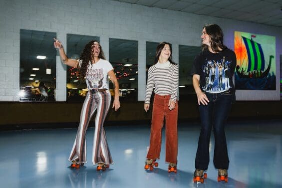 Three women roller skating at Viking Skate Country in Redding, CA, with colorful retro pants and roller skates on a blue rink floor. A colorful Viking ship painting is visible on the wall in the background.